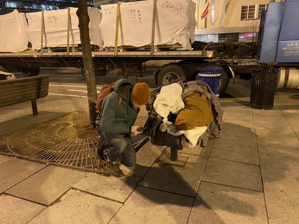 A man kneeling down beside a homeless person a bench, asking them some questions