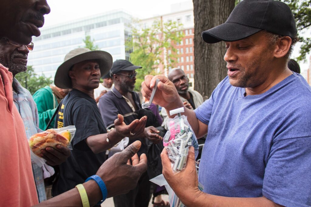 A street outreach volunteer passes disposable razors to a group of men.
