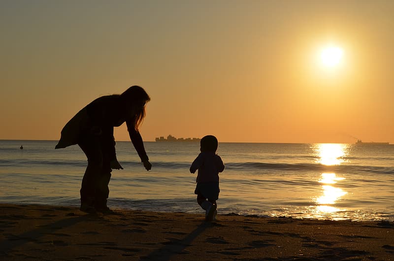 A woman reaching out for her baby on the beach. Sunset in the background.