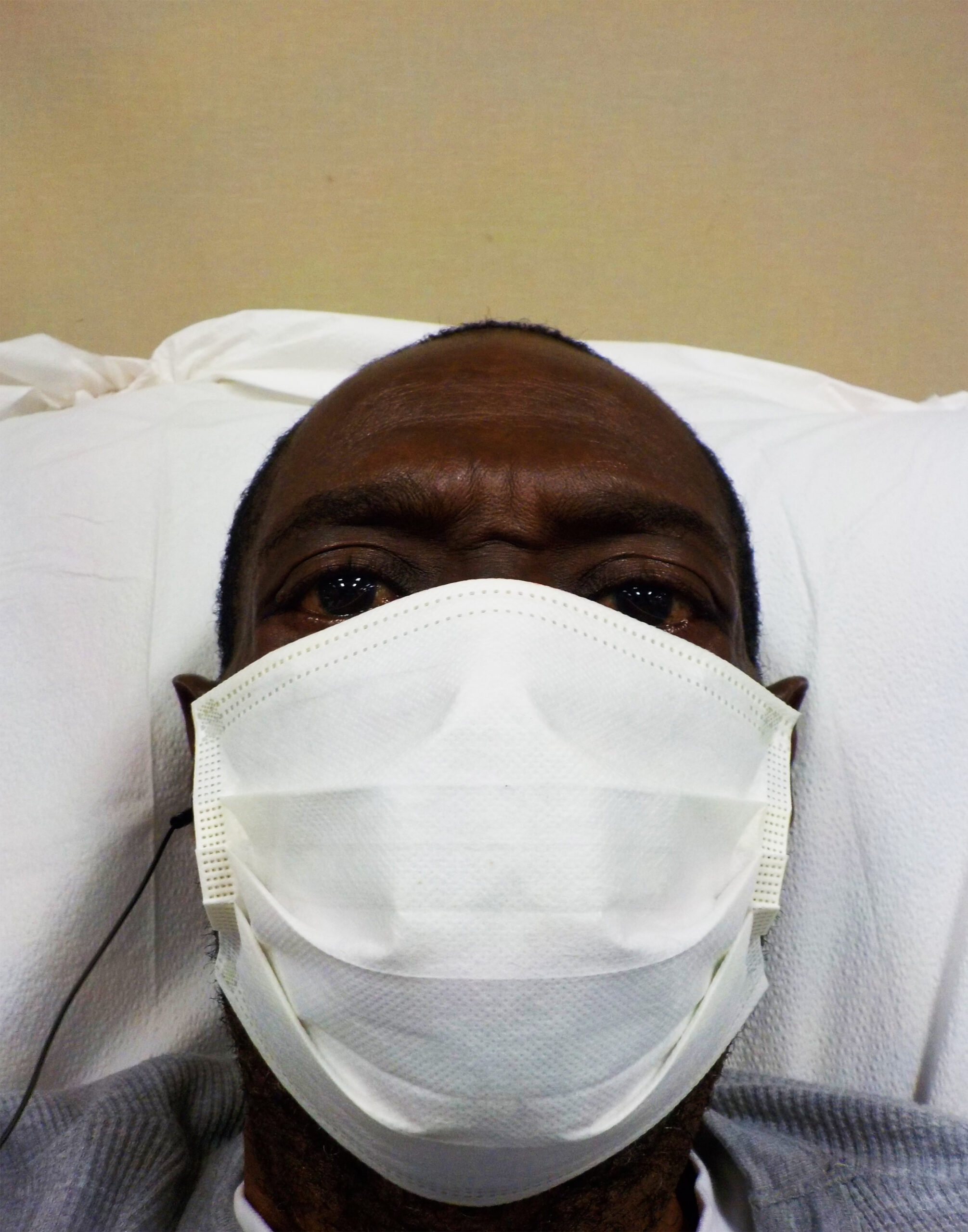 Photo showing Joseph's head, laying on a pillow in a medical facility, wearing a mask.