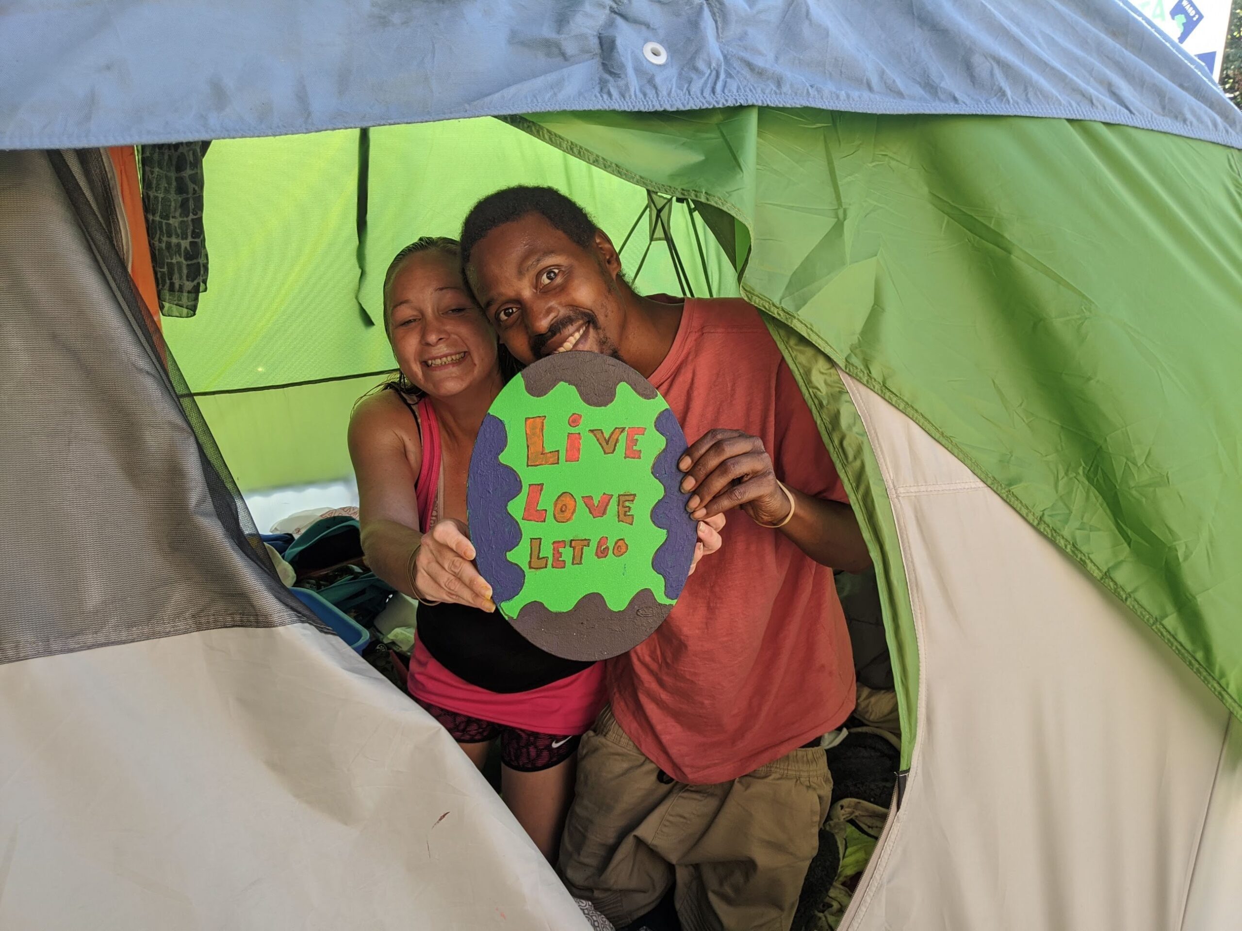 Picture of a woman and a man holding up a hand-painted sign that says "Live Love Let Go" as they kneel inside of a green and gray tent with the entry flap open.