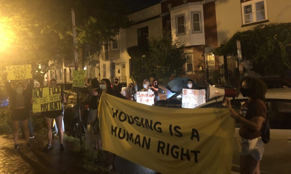 Protesters hold a yellow banner saying "Housing is a Human Right" and signs on the sidewalk.
