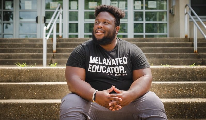 Photo showing a man smiling with his hands clasped, sitting on steps in front of a school building, wearing a shirt that says "Melanated Educator."