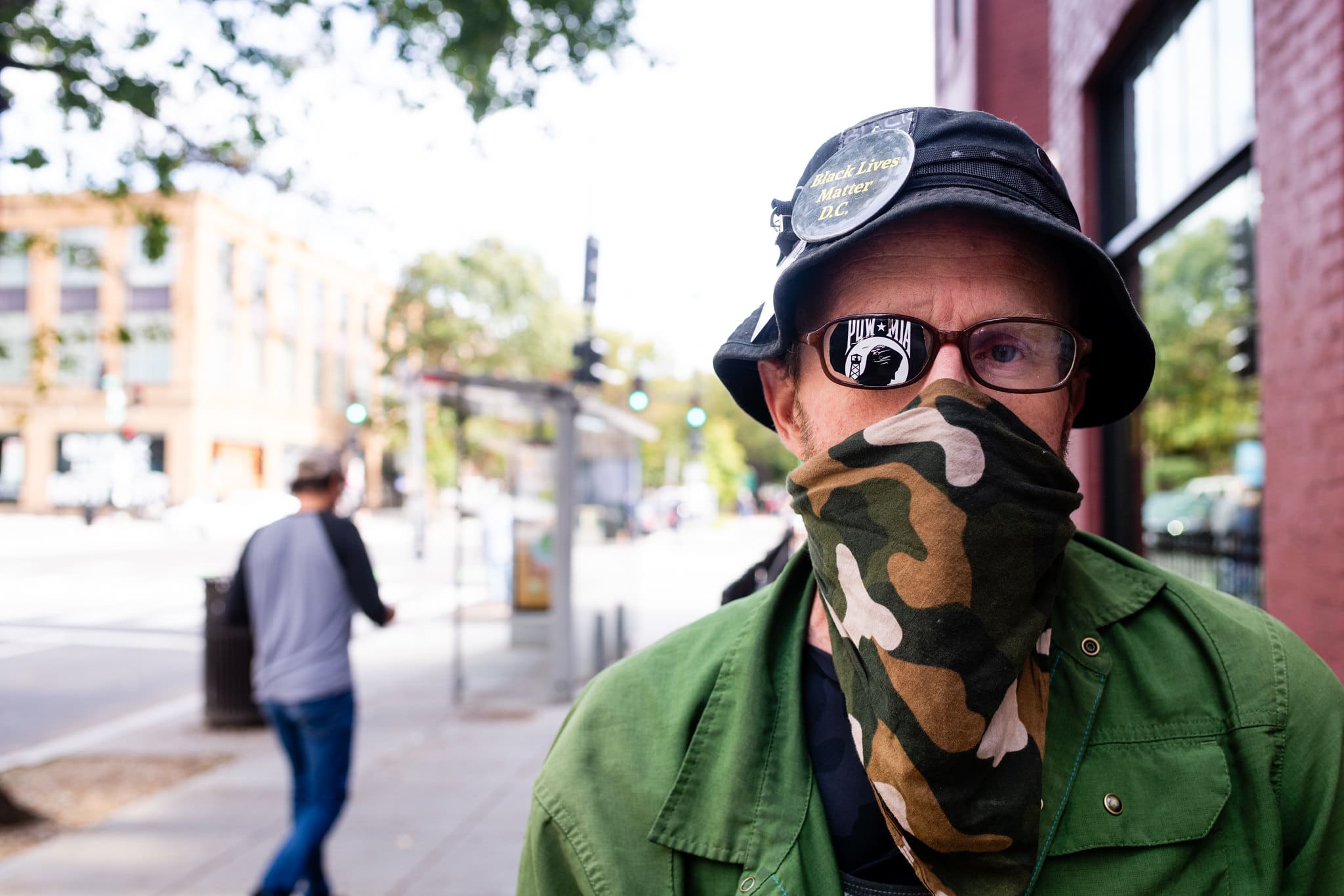 Close-up photo of Saul from the shoulders up. A bright streetscape is visible behind him. He's wearing a black bucket hat with a button that says "Black Lives Matter" along with a "Prisoner of War" flag eyepatch and using a camouflage-print bandana as a face mask.