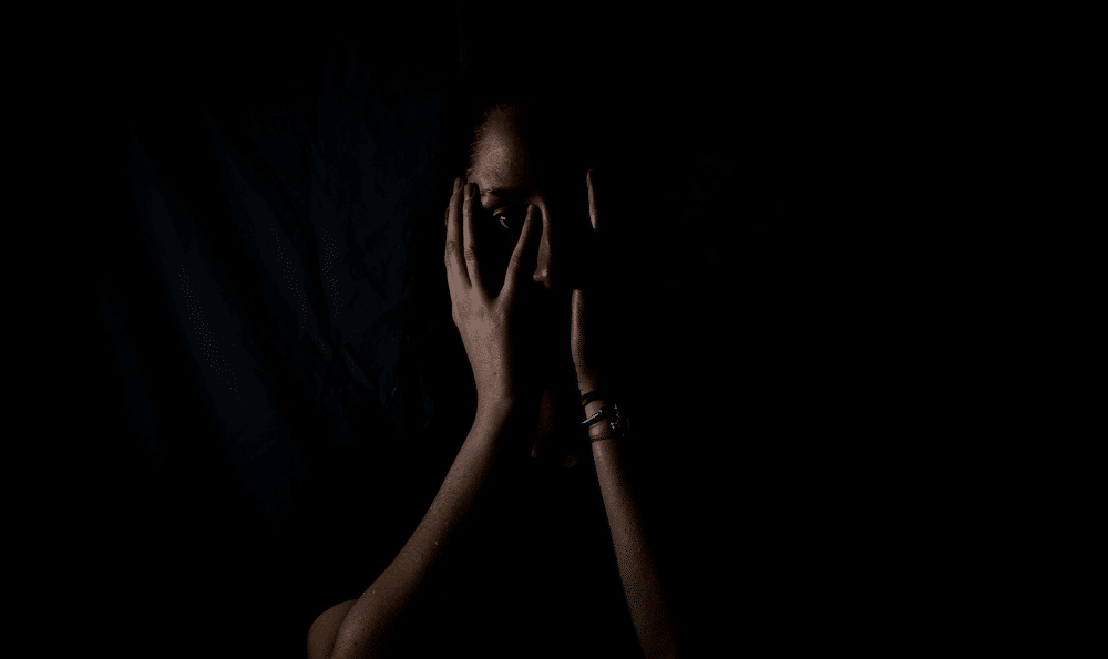 Photo of a woman with dark skin, mostly hidden in shadow, covering her face with her hands.