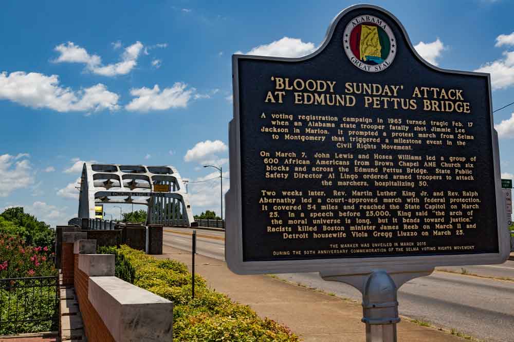 Photo of a sign about "Bloody Sunday" in the foreground and the Edmund Pettus Bridge in the background, taken on a sunny day.