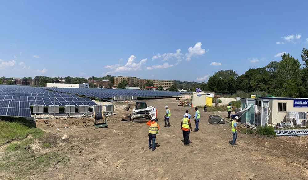 Photo showing several construction workers installing many solar panels in a mostly dirt field that is surrounded by trees with city buildings visible in the distance.