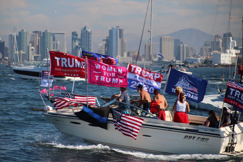 A boat holding people waving different flags, predominantly American and Trump flags.