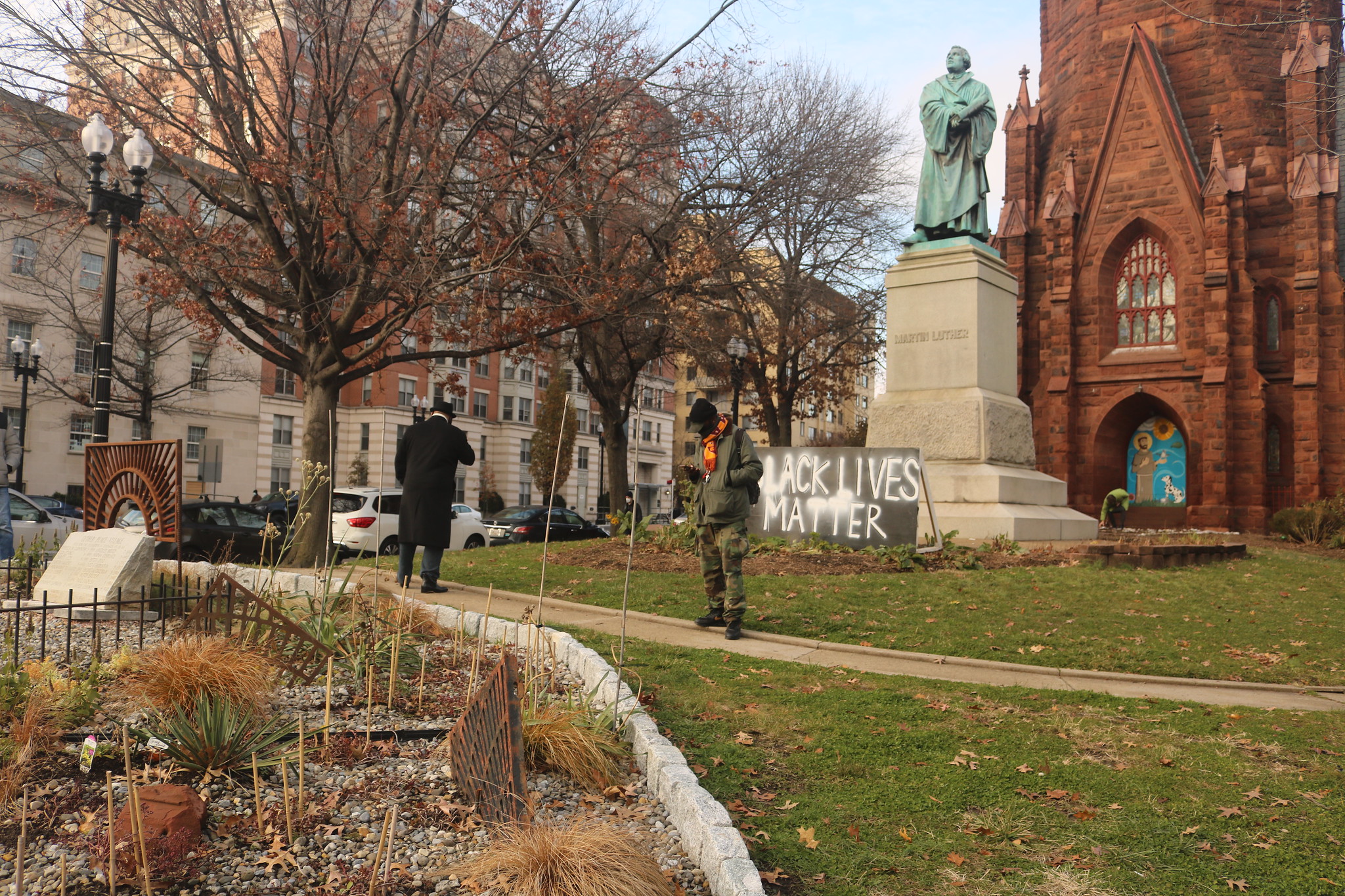 Photo of a garden, two people standing on a path in the grass, and a statue of Martin Luther with a "Black LIves Matter" sign leaning against it in front of a red church building.