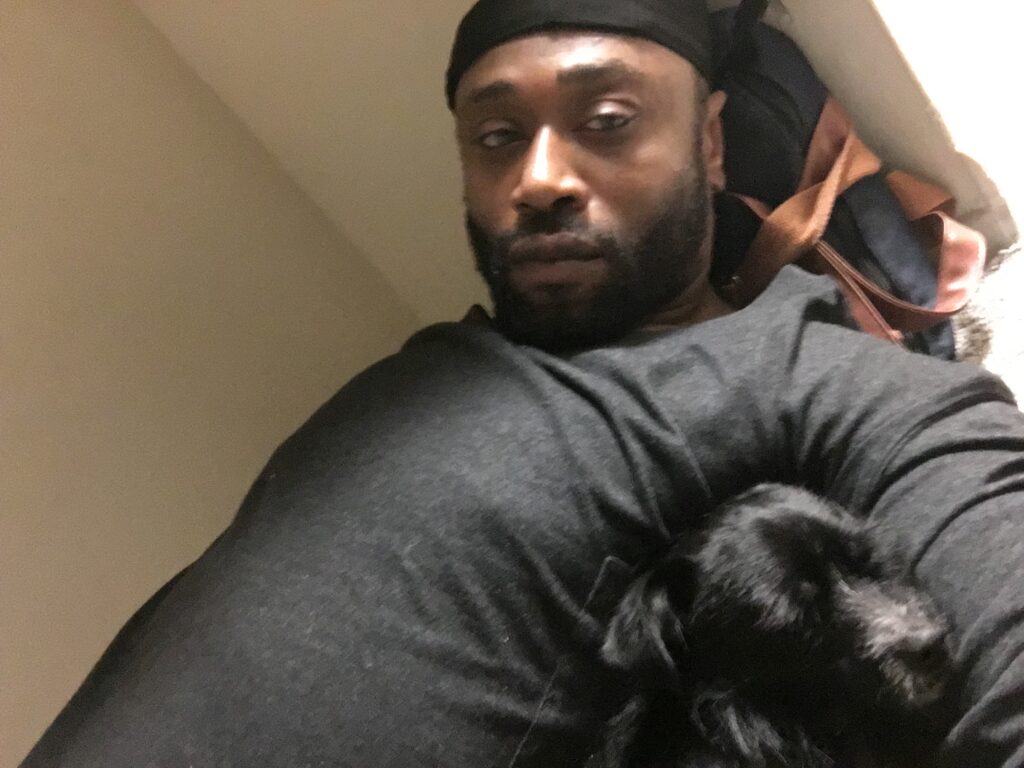 A Black man in a black shirt takes a picture with his dog.