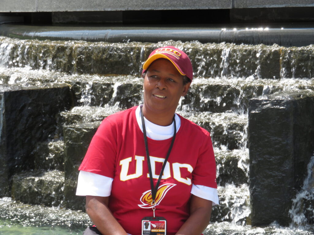 Photo of a woman in a red and white "UDC" shirt and hat, wearing a student ID around her neck, sitting in front of a waterfall on the UDC campus.