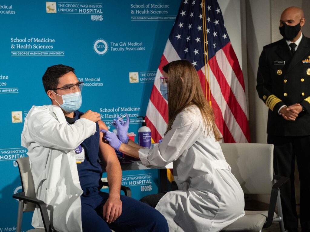 A man is getting a vaccine shot in the arm by a doctor. Both are wearing masks and are sitting in front with George Washington University Hospital labels.