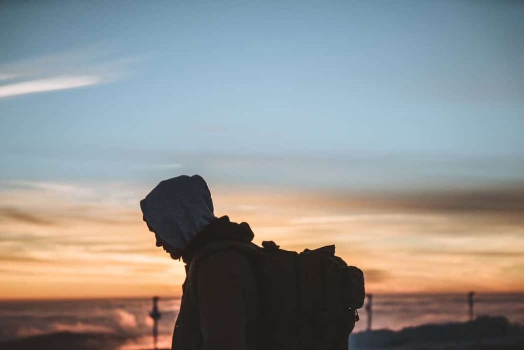 A man wearing a backpack and warm clothing stands in front of sunset.
