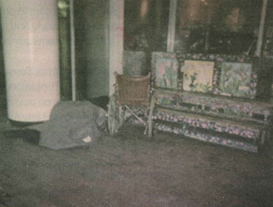 Photo of person laying beneath a blanket on the sidewalk, next to an empty wheelchair.