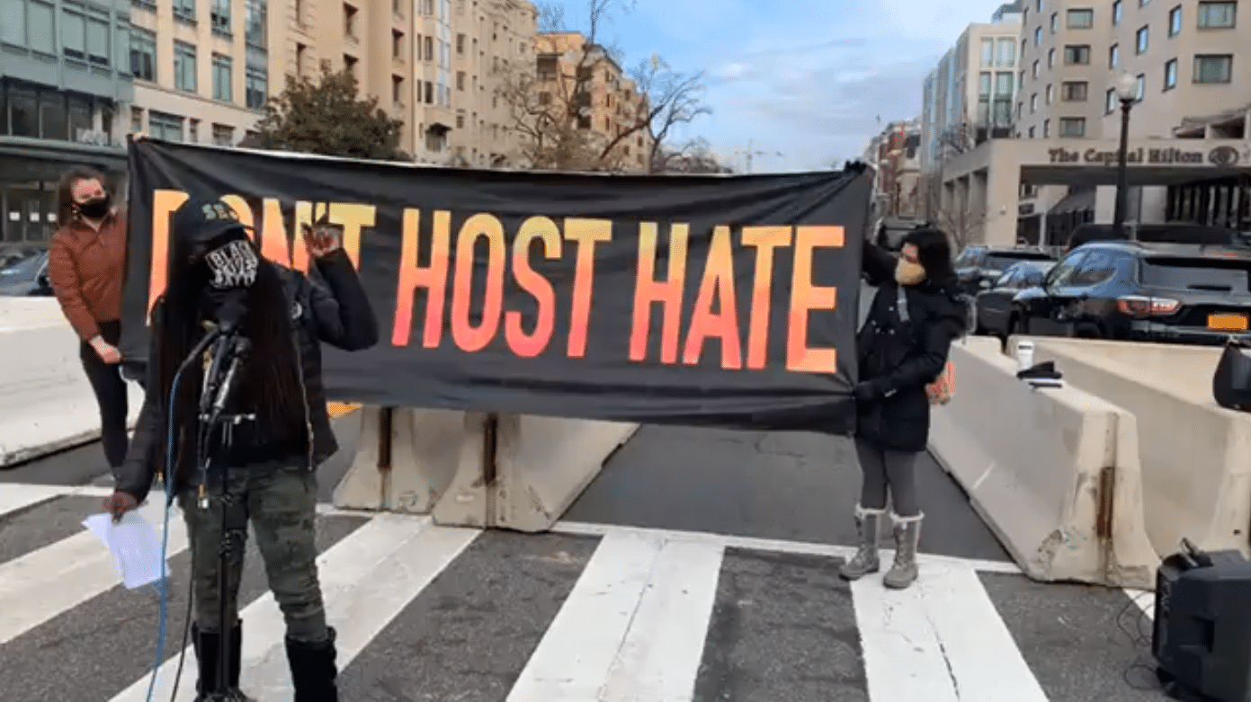 A woman in a "Black Lives Matter" mask speaks while two other people hold up a banner behind her that says "DON'T HOST HATE." The Capitol Hilton hotel can be seen in the back-right corner.