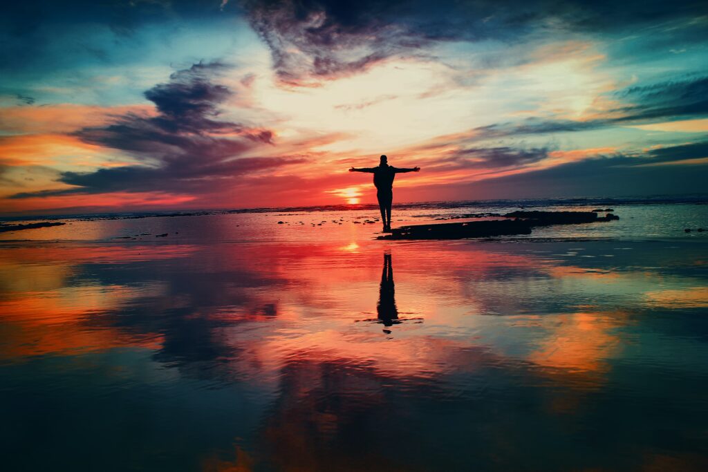 Photo shows a person standing with their arms open wide with a pink and blue sunset behind them