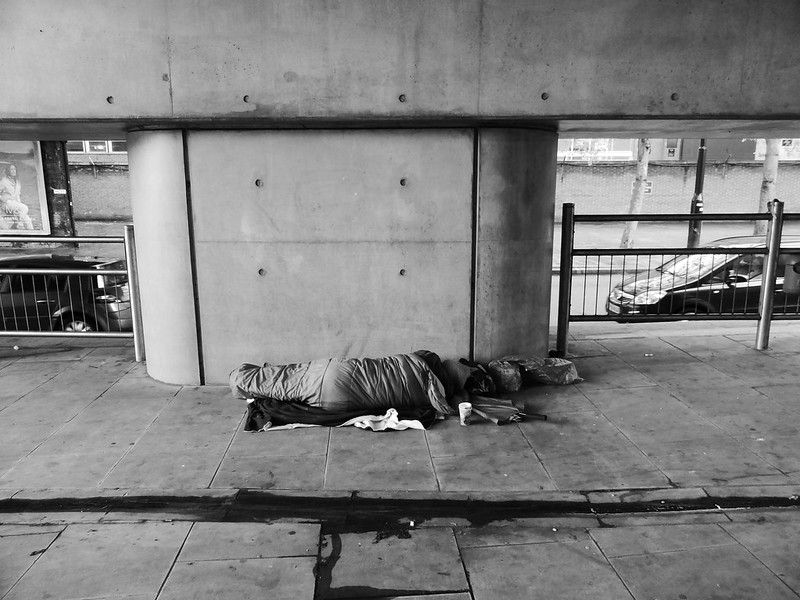 A black and white photo of a man sleeping on the street, his face is not shown and he is sleeping in a sleeping bag