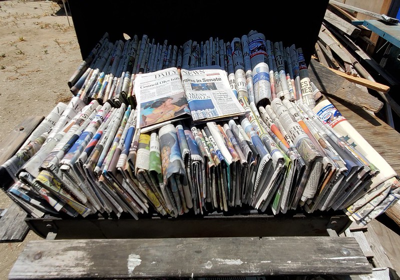 A photo of newspaper piles., with two papers folded out to say "Daily News"