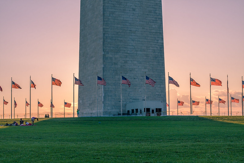 Photo of the base of the Washington Monument at sunset. Numerous American Flags are waving in the wind and a family is picnicking.