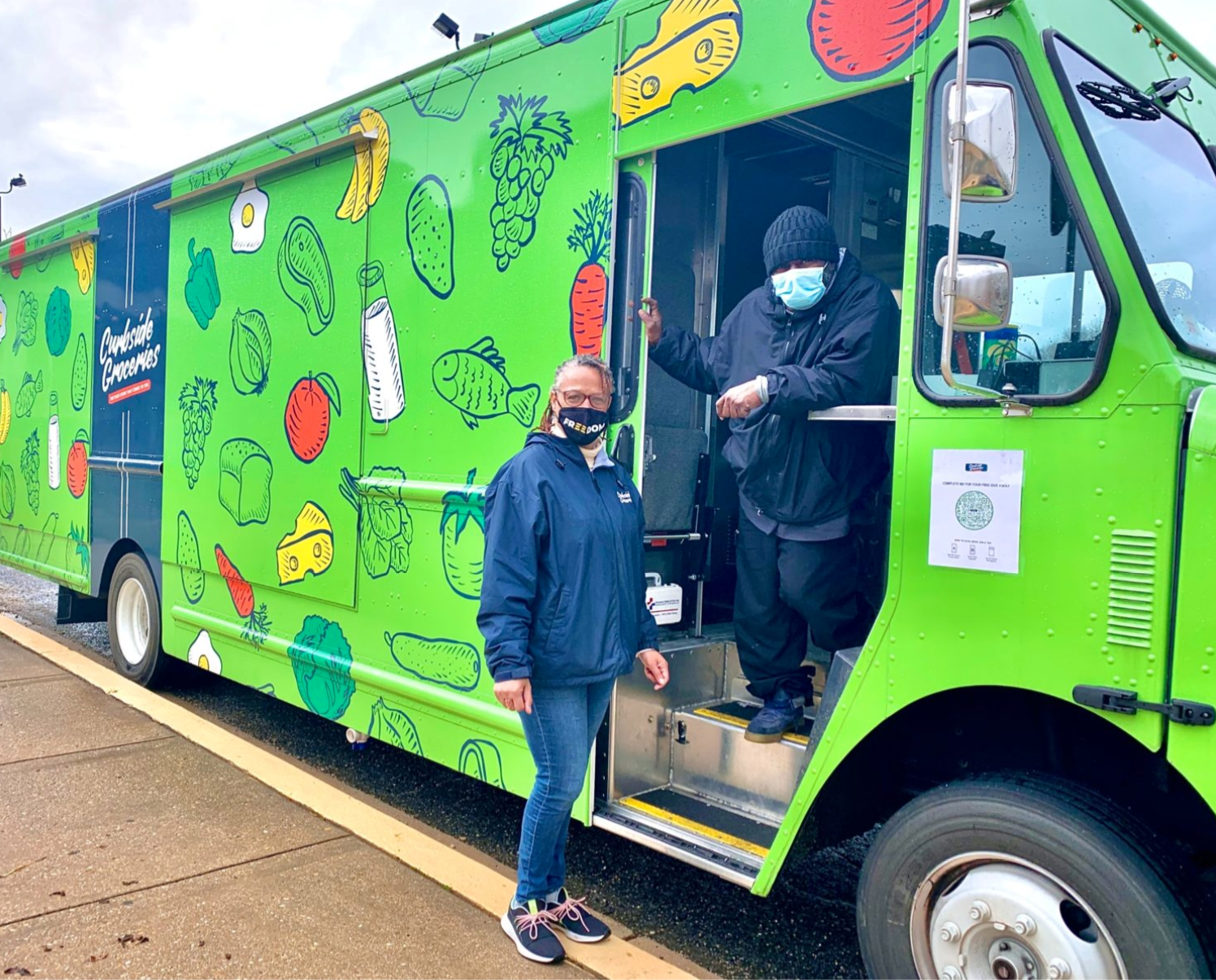 Photo displays two workers outside of a bright green food ruck with cartoon vegetables on the truck