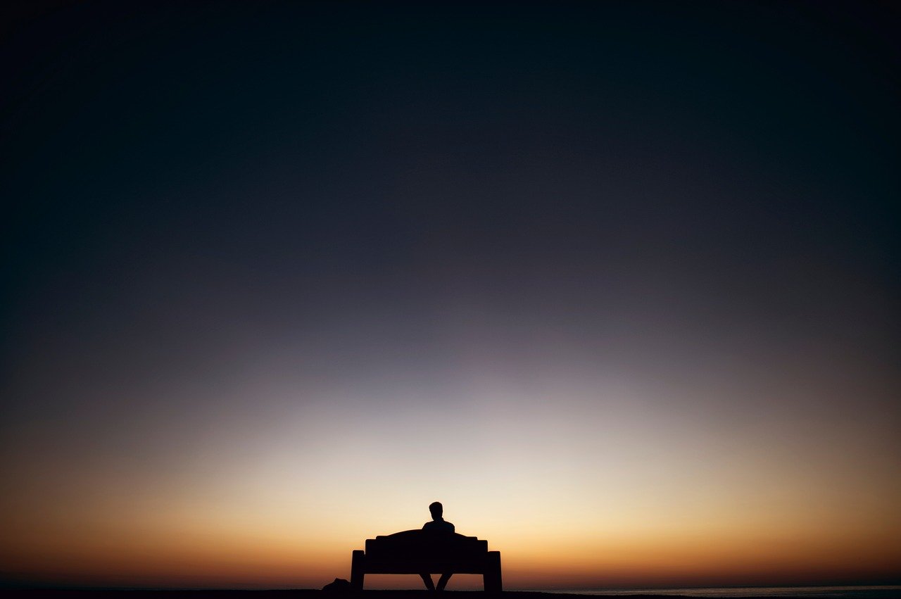 The shadowed outline of a man sitting by himself on a bench.