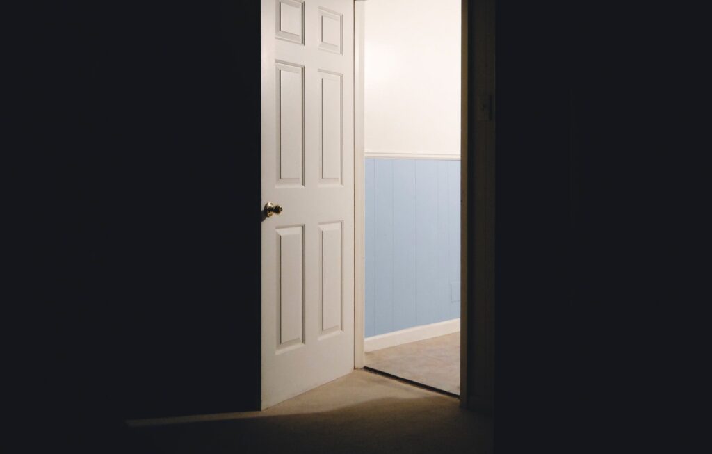 Photo showing an open door in a dark room. A lit hallway is visible through the opening.
