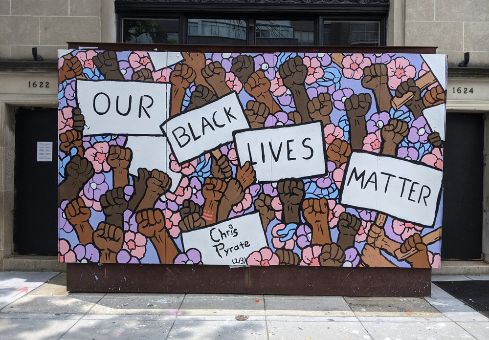 Many dark-skinned hands on a brightly colored background. Some hold individual signs that bear single words. Together they read "OUR BLACK LIVES MATTER"