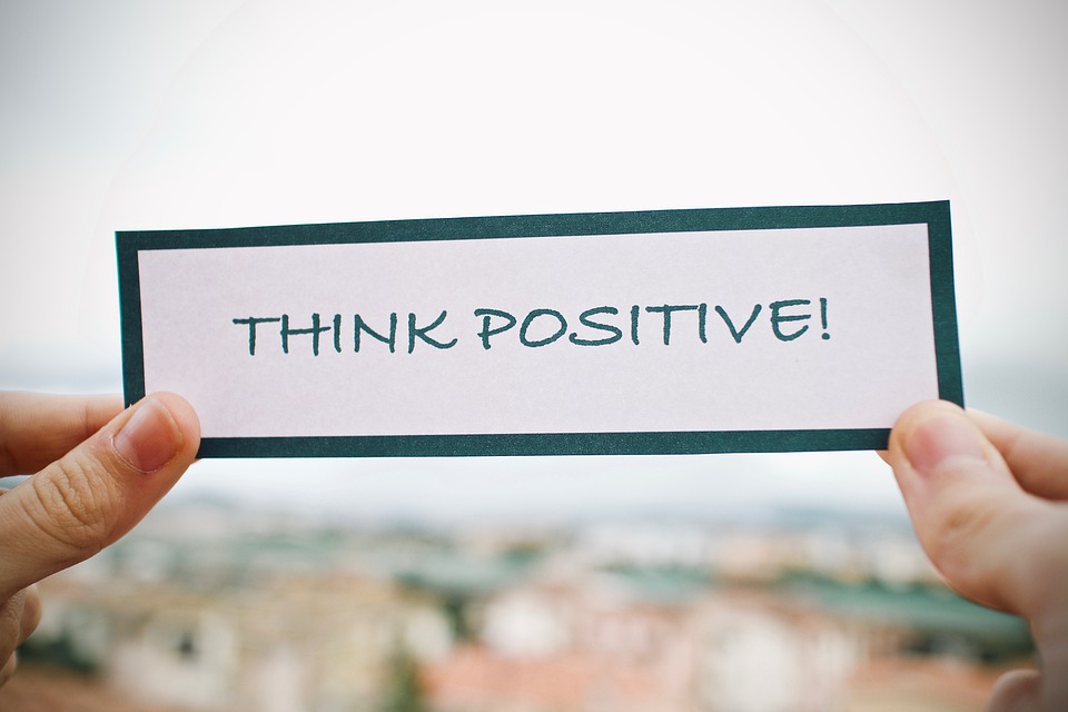 Photo of hands holding up a "Think Positive!" paper sign in front of a blurred out view
