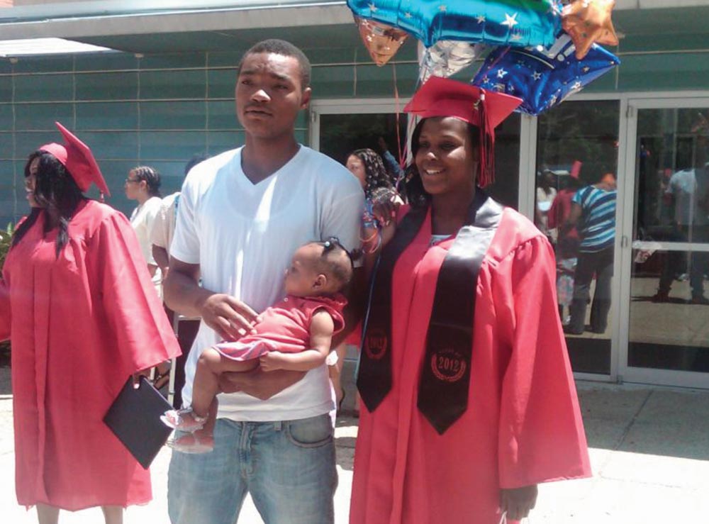 Photo of a young woman in a graduation cap and gown and holding balloons, standing with a young man holding a child.