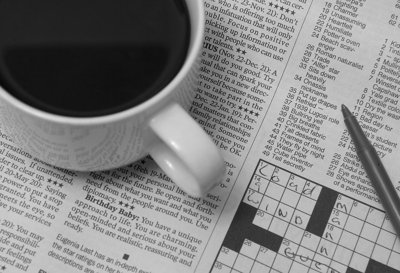 A cup of coffee sitting on a newspaper opened to the crossword puzzle.