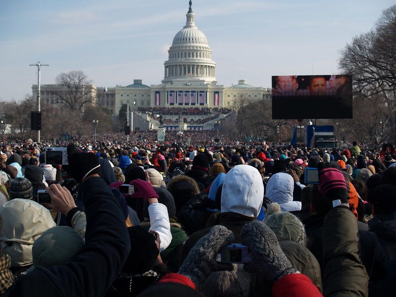 A photo of a large crowd gathered to watch the Inauguration. The Capitol and a jumbotron showing President Obama are in the background.