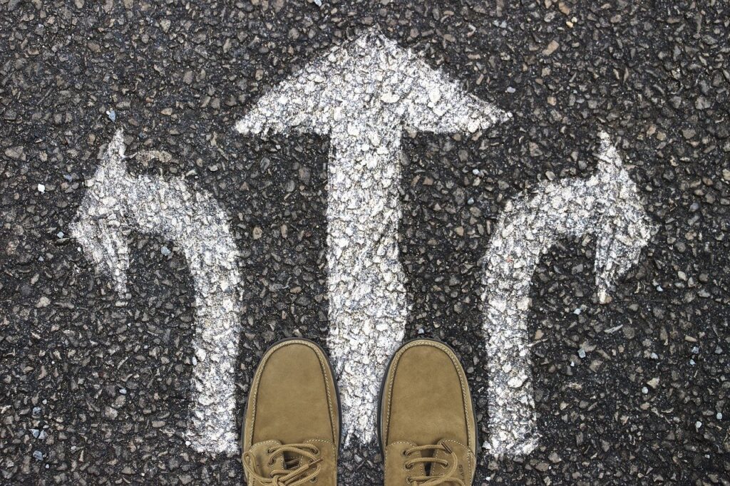 Photo of a person standing at a crossroads of arrows. Only the tip of the person's shoes are visible. The arrows are painted on the ground.