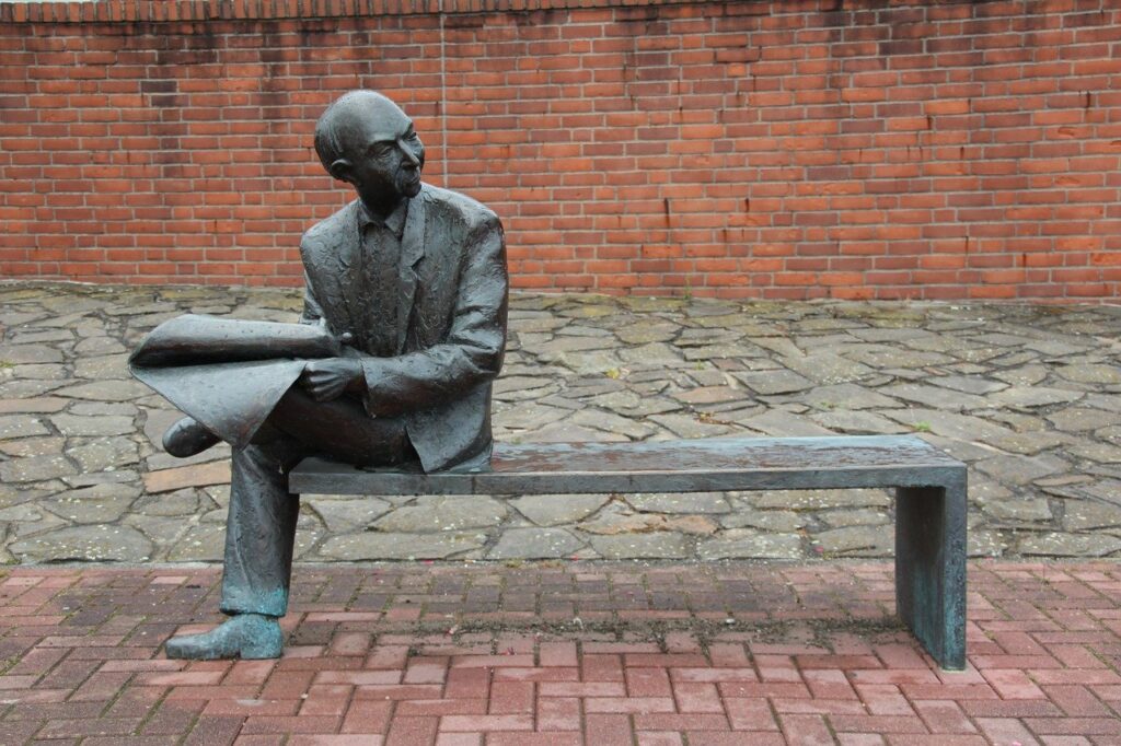 Photo of a statute of a man sitting on a bench, reading a newspaper.
