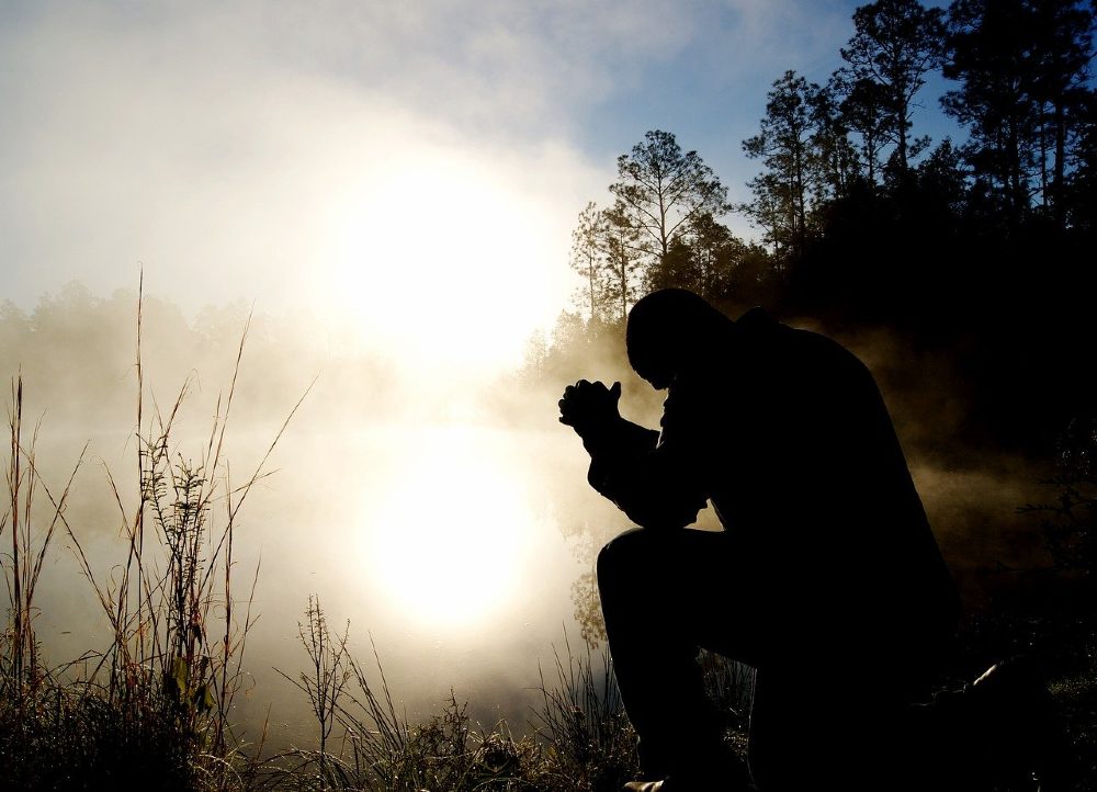 Photo of a man praying bended knees outside in nature during a foggy sunrise.