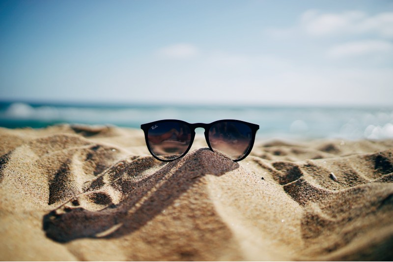 Photo of a pair of sunglasses sitting in sand on a beach; the ocean is in the background