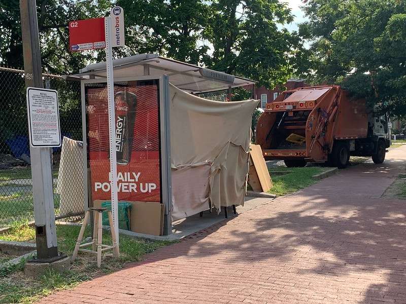 Photo of a garbage truck parked next to a bus shelter that has been converted into a shelter for unhoused residents using blankets.