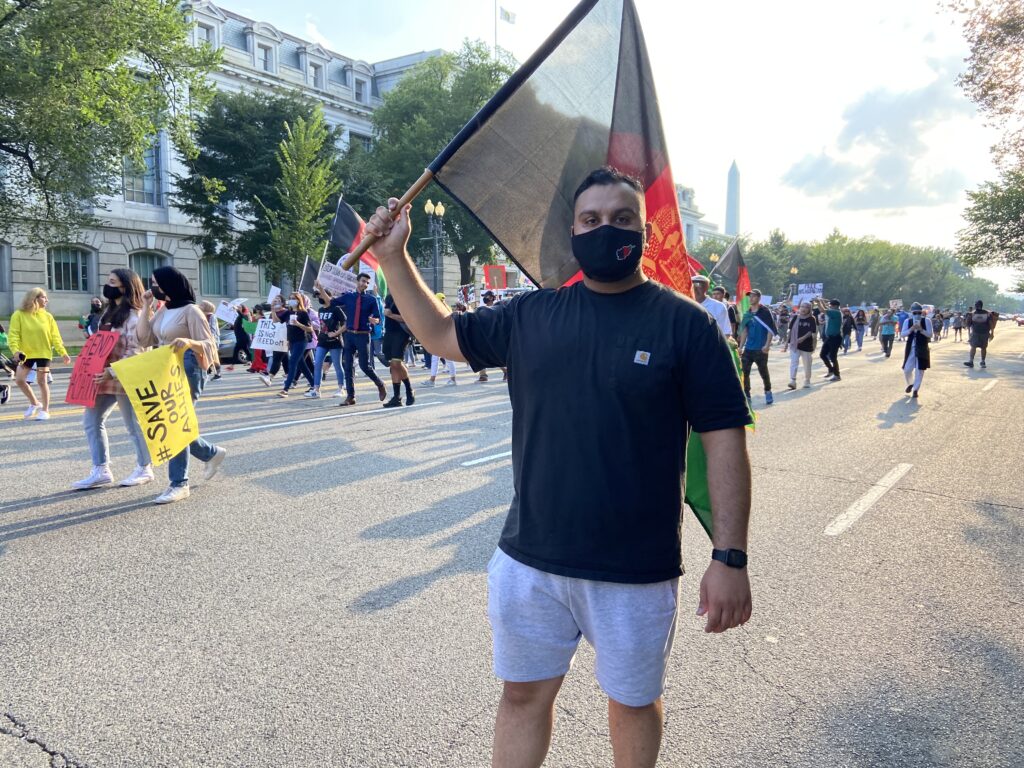 A man marches in a crowd on a street in DC holding a large black, red, and green Afghanistan flag.