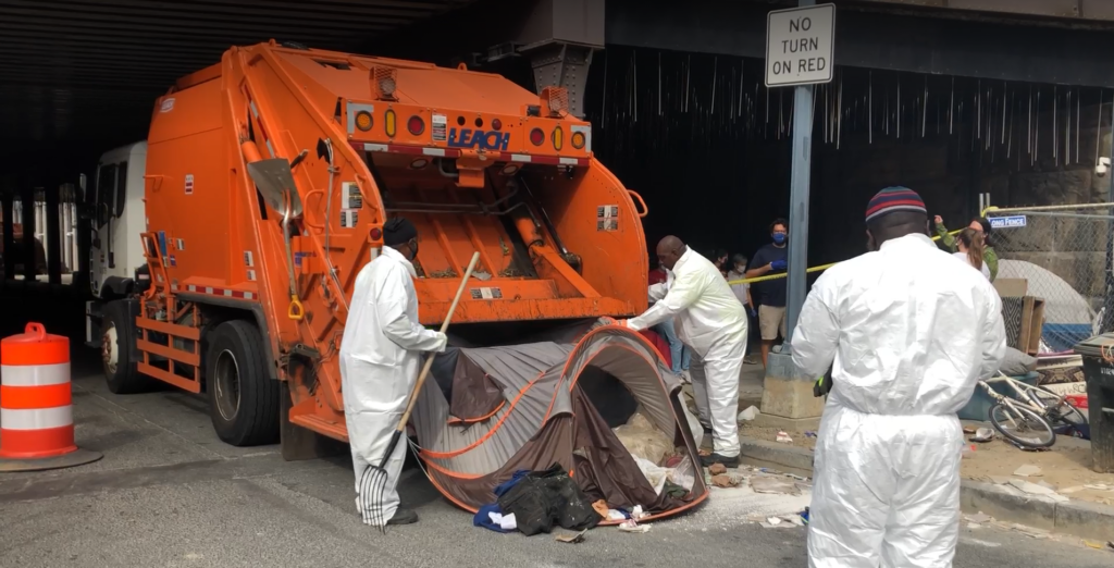 Three waste collectors prepare to load a homeless tent into hopper of a garbage truck