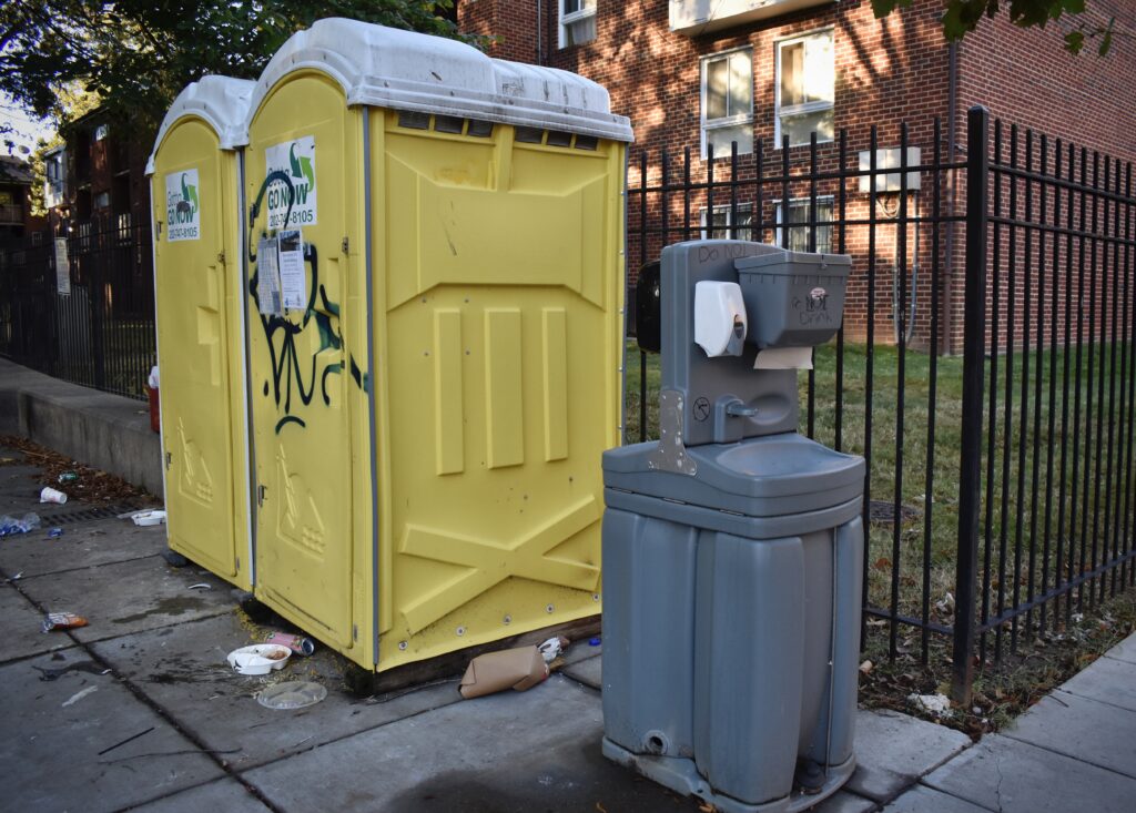 Two porta potties placed against a black fence with a handwashing station