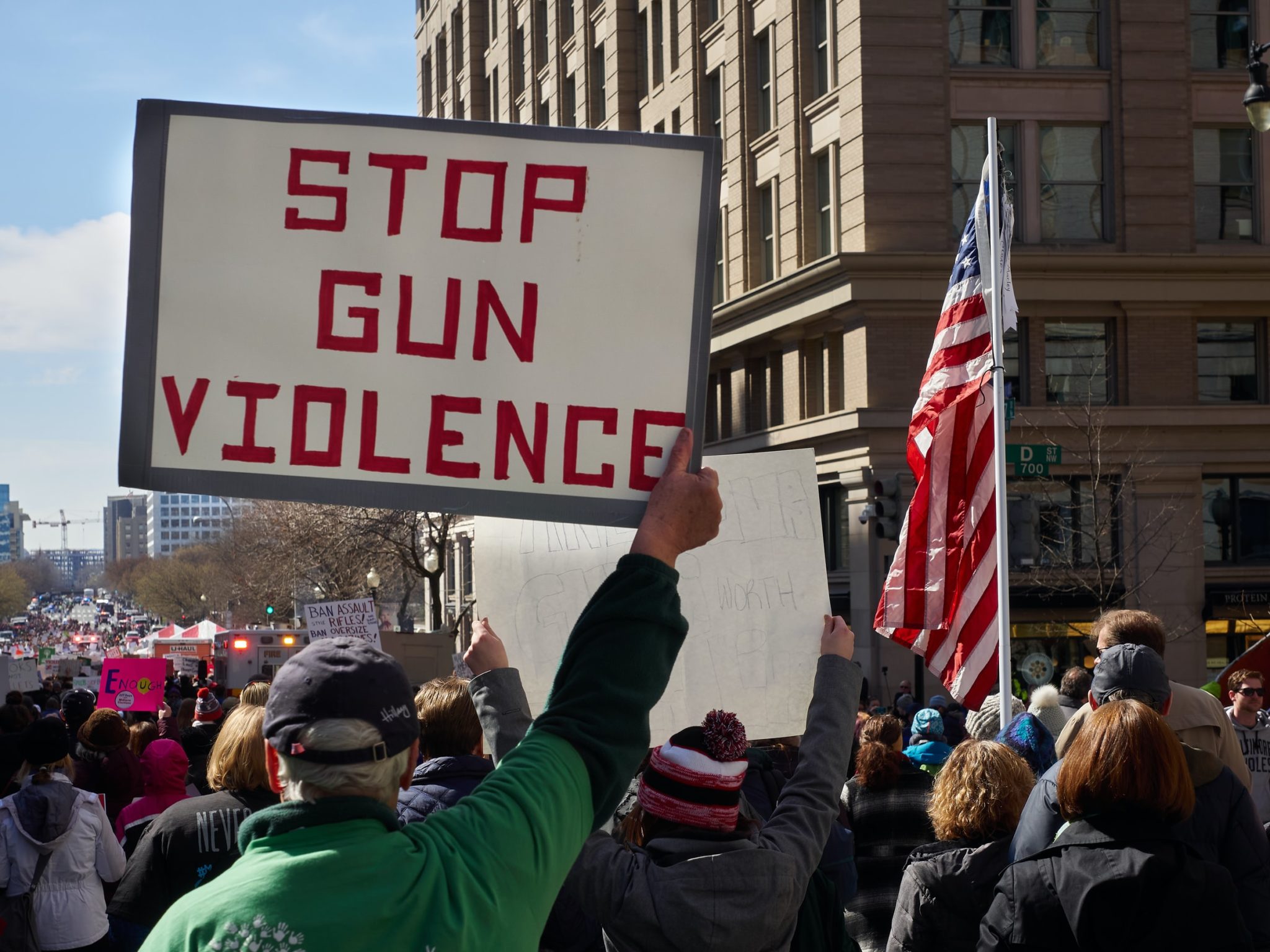 Crowds of protestors in the street. One holds a sign that says "Stop Gun Violence." Another holds an American flag.