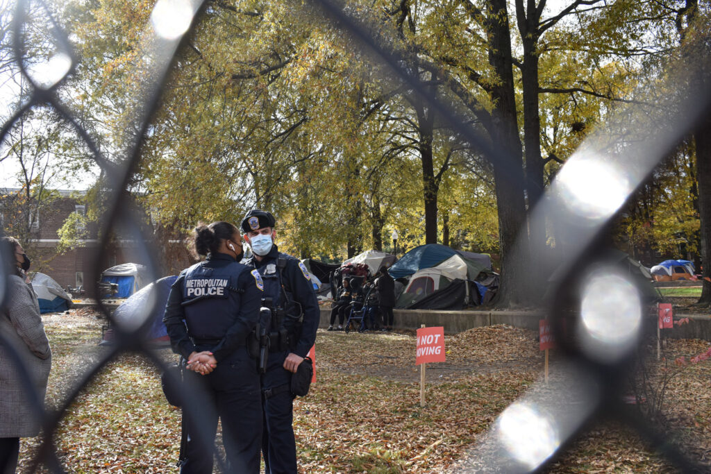 Two officers stand in the encampment. While the black metaling fencing acts like a boarder around the two officers.