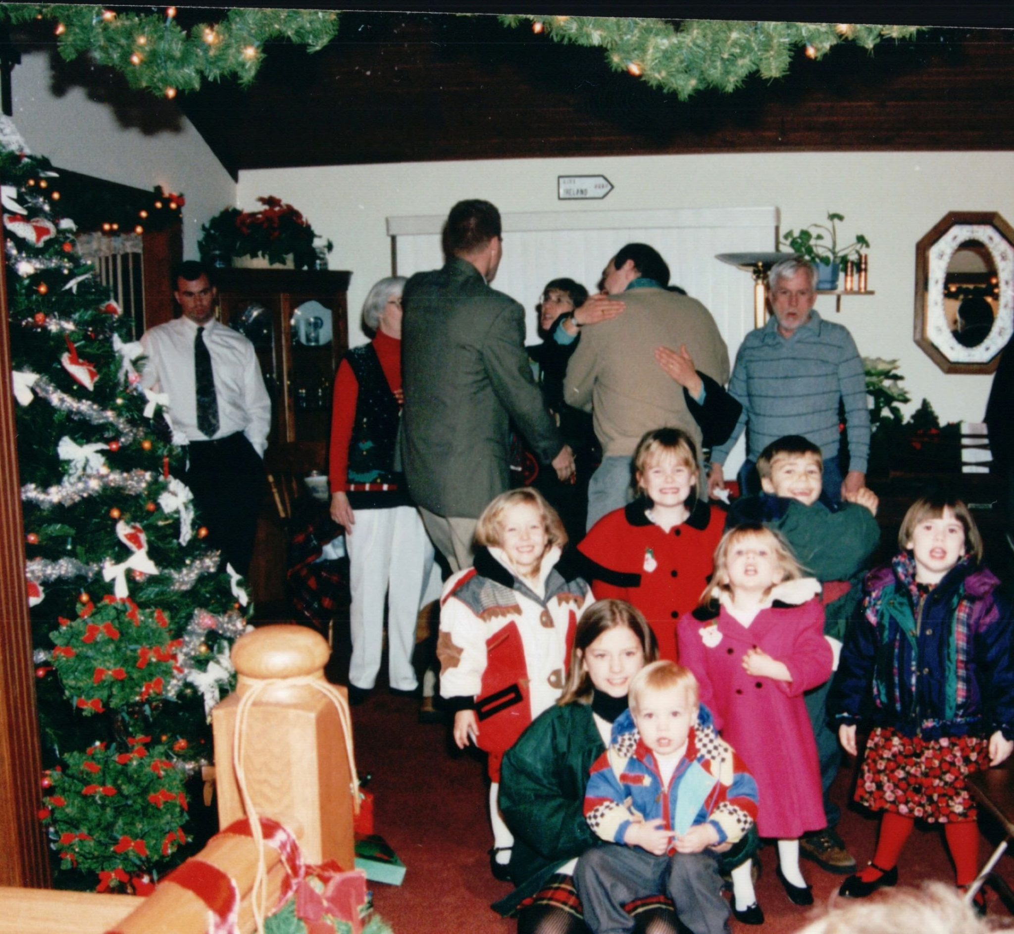Photo of the Daily family Christmas. To the left a Christmas tree decorated in tinsel with adults embracing in the background. In the foreground 7 kids pose for the picture and to the right Bill Daily, in a blue shirt, walks toward the camera.