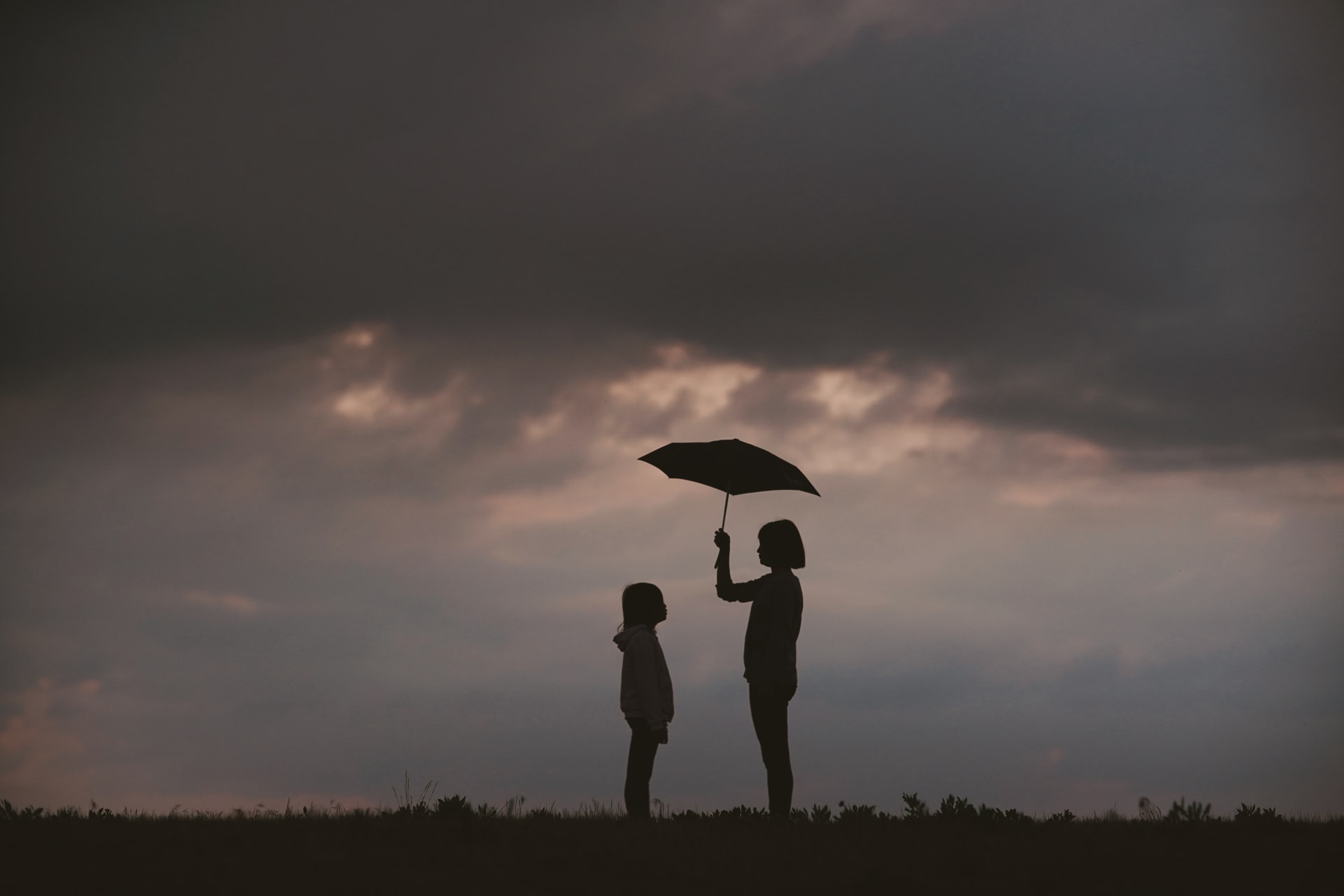 Photo of an taller person protecting a shorter person against the rain with an umbrella. The figures are silhouetted against a grey backdrop.