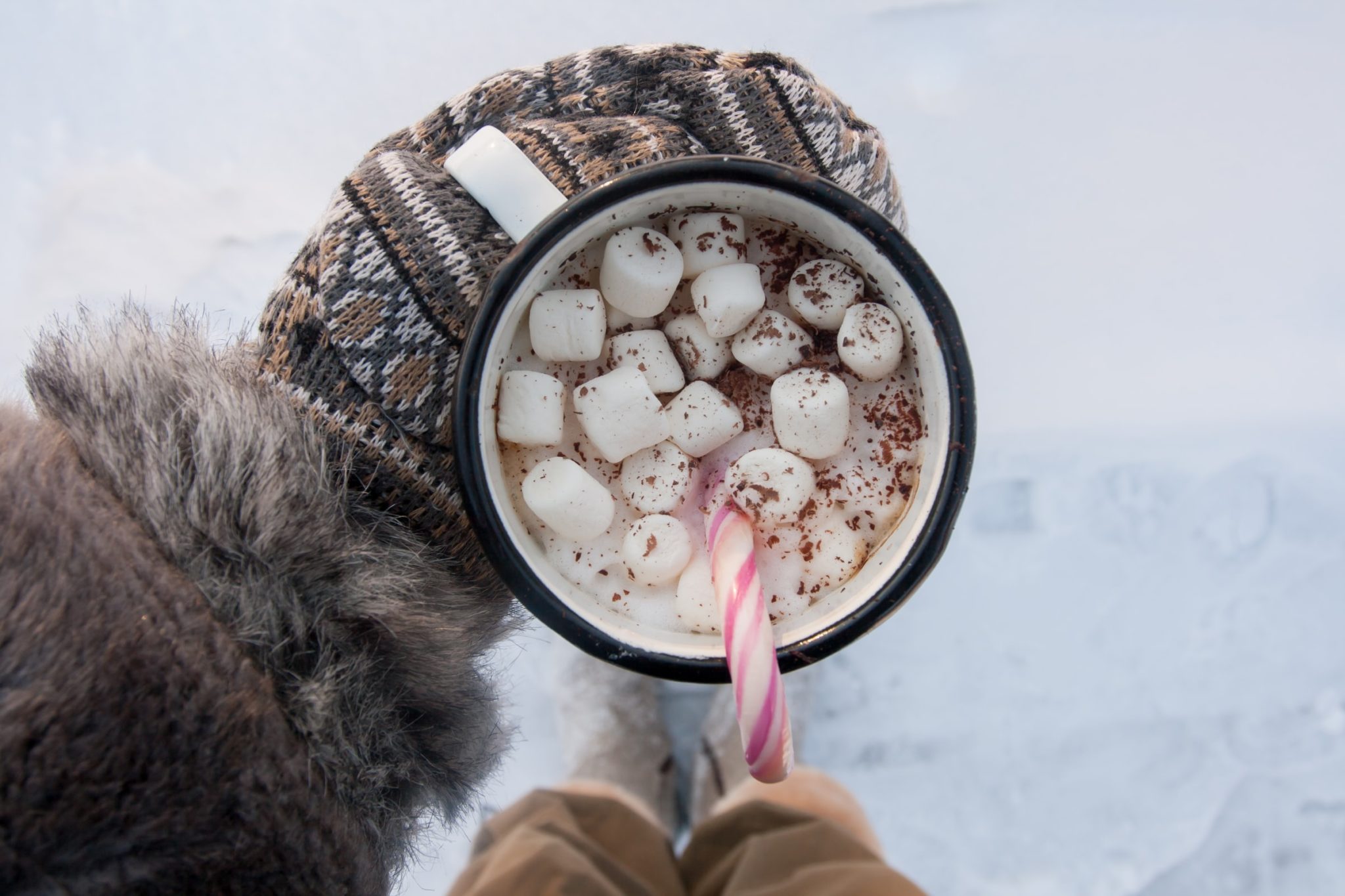 Photo of a cup of hot chocolate with marshmallows and a candy cane being held by a hand in a brown and white mitten.