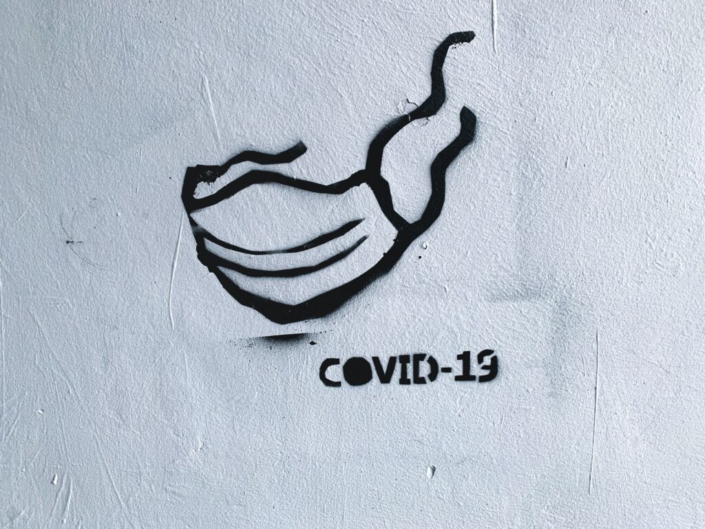 Photo of an outline of a mask spray painted on a grey wall. To the bottom left COVID -19 is also spray painted on the wall in black.