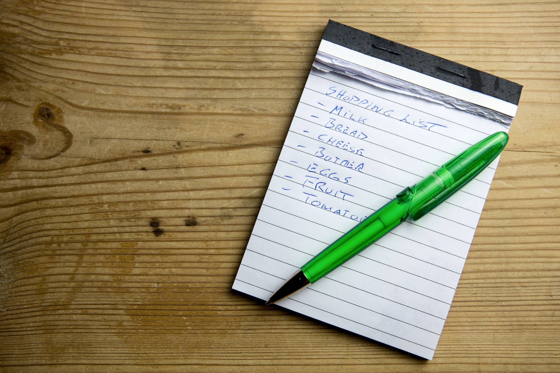 A photo of a shopping list written on a note pad with a green pen laid on top of the notepad.