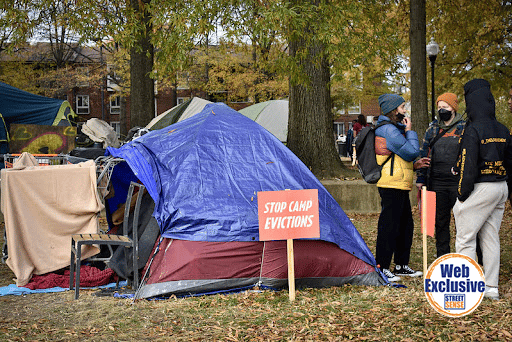 Photo of tents with a sign that reads "Stop camp evictions." Also, a group of people in winter gear have a conversation to the right of the photo