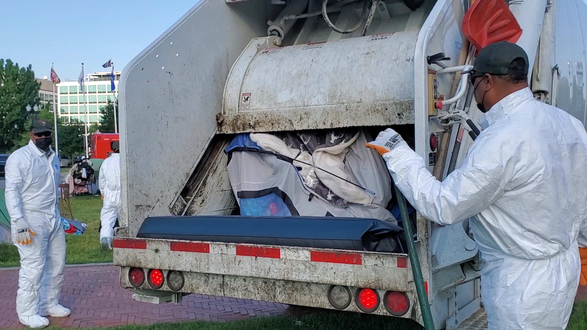 Two people in white protective suits look at a trash truck filled with tents.