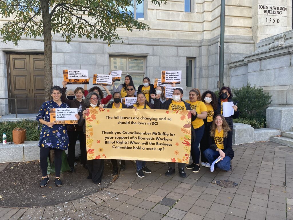 A group of people in yellow shirts stand behind a banner that calls on the D.C. Council to pass the domestic workers bill of rights.