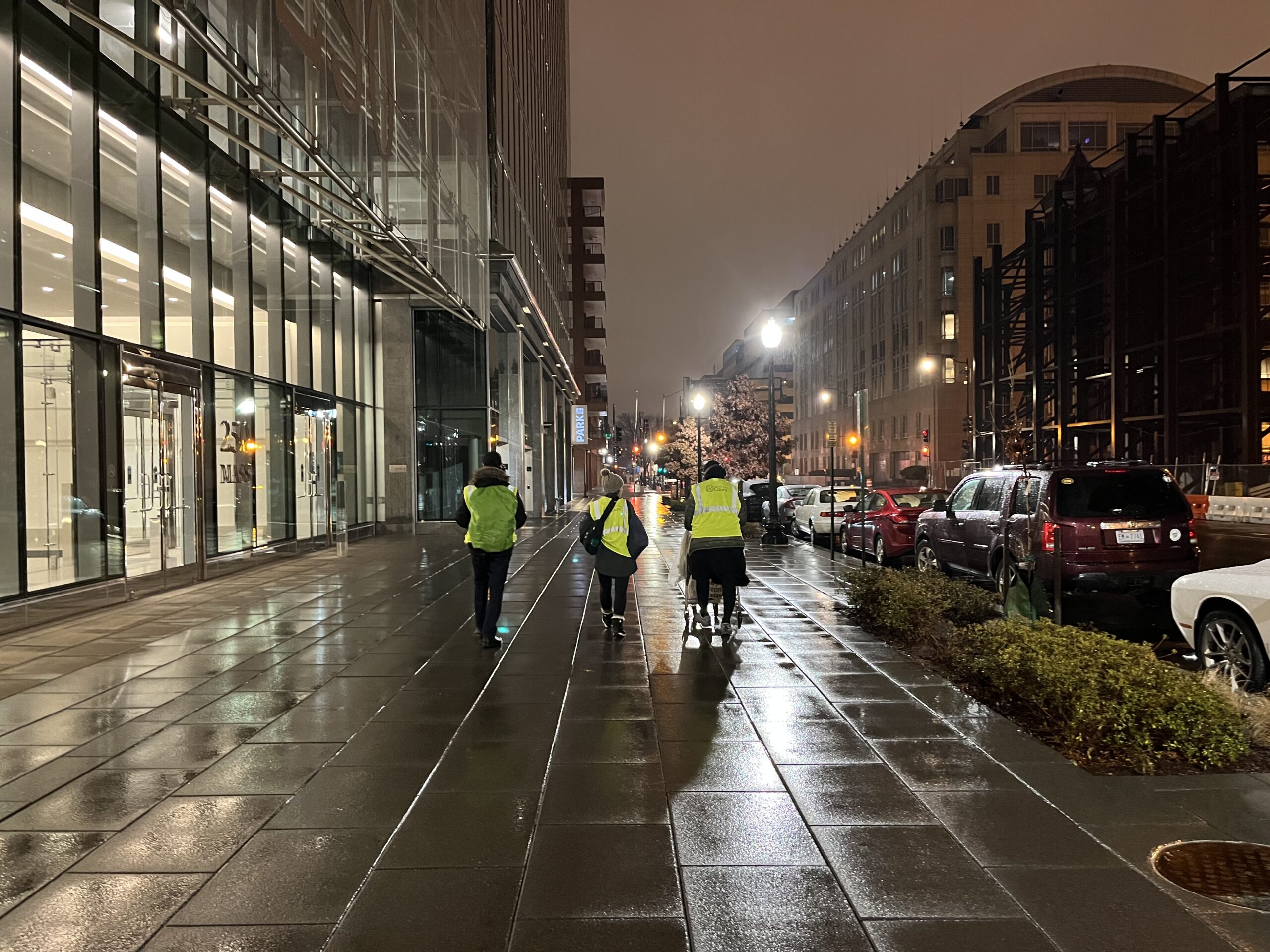 Three people in yellow vests walk down a rainy street looking for people experiencing homelessness.